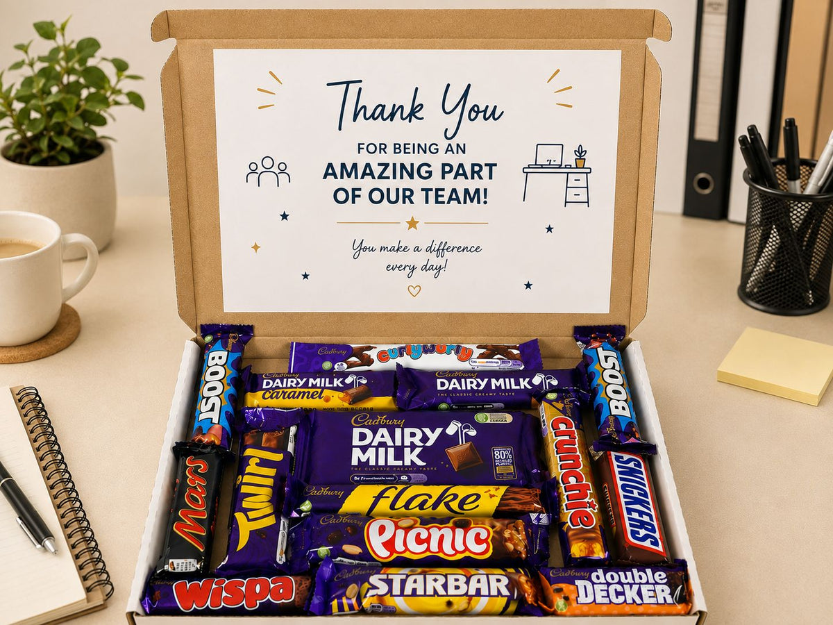 An open cardboard gift box on a desk filled with assorted chocolate bars, including Cadbury Dairy Milk, Flakes, Snickers Twix, Mars, and Decker. The box lid has a printed thank-you card that reads 'Thank You FOR BEING AN AMAZING PART OF OUR TEAM!'.