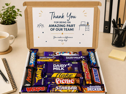 An open cardboard gift box on a desk filled with assorted chocolate bars, including Cadbury Dairy Milk, Flakes, Snickers Twix, Mars, and Decker. The box lid has a printed thank-you card that reads 'Thank You FOR BEING AN AMAZING PART OF OUR TEAM!'.