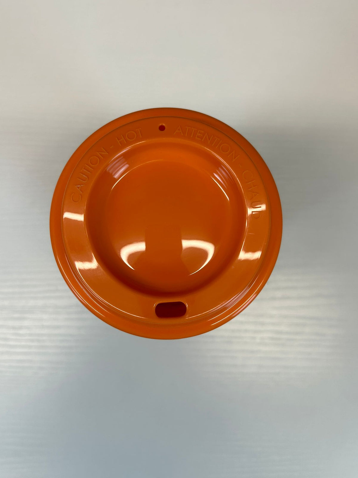 An orange plastic lid with a circular design, featuring embossed text reading 'CAUTION HOT' around the rim and a small cutout near the bottom, shown from a top-down view 